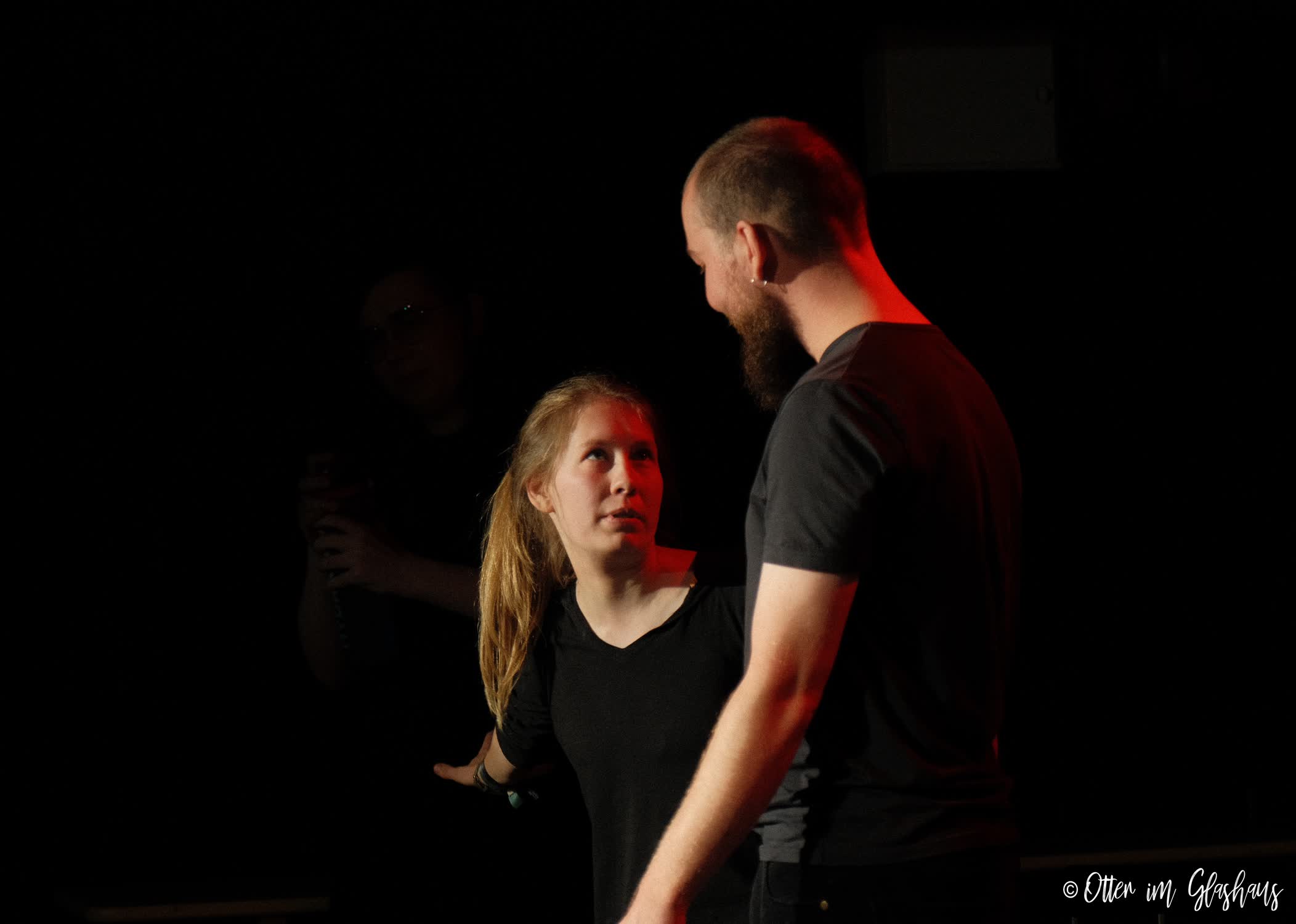 Zwei Schauspielende. Die eine schaut den anderen flehend an. Rotes licht scheint ihr ins Gesicht.