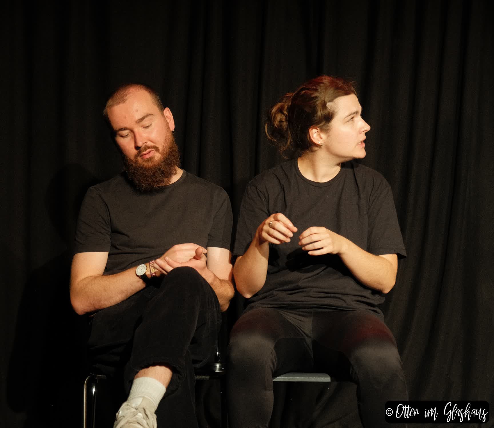 Zwei Schauspielende sitzen auf Stühlen. Einer schaut auf seine Hand, die andere blickt interessiert zur Seite.